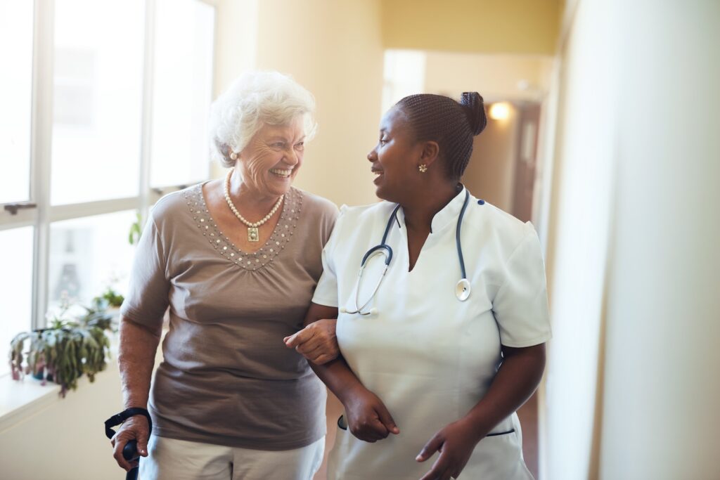 Smiling nurse walks arm-in-arm with an older woman in a hallway, both laughing warmly.
