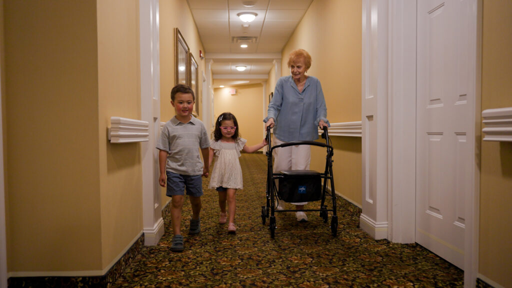 Older woman using a walker walks alongside two children in an assisted living community hallway.